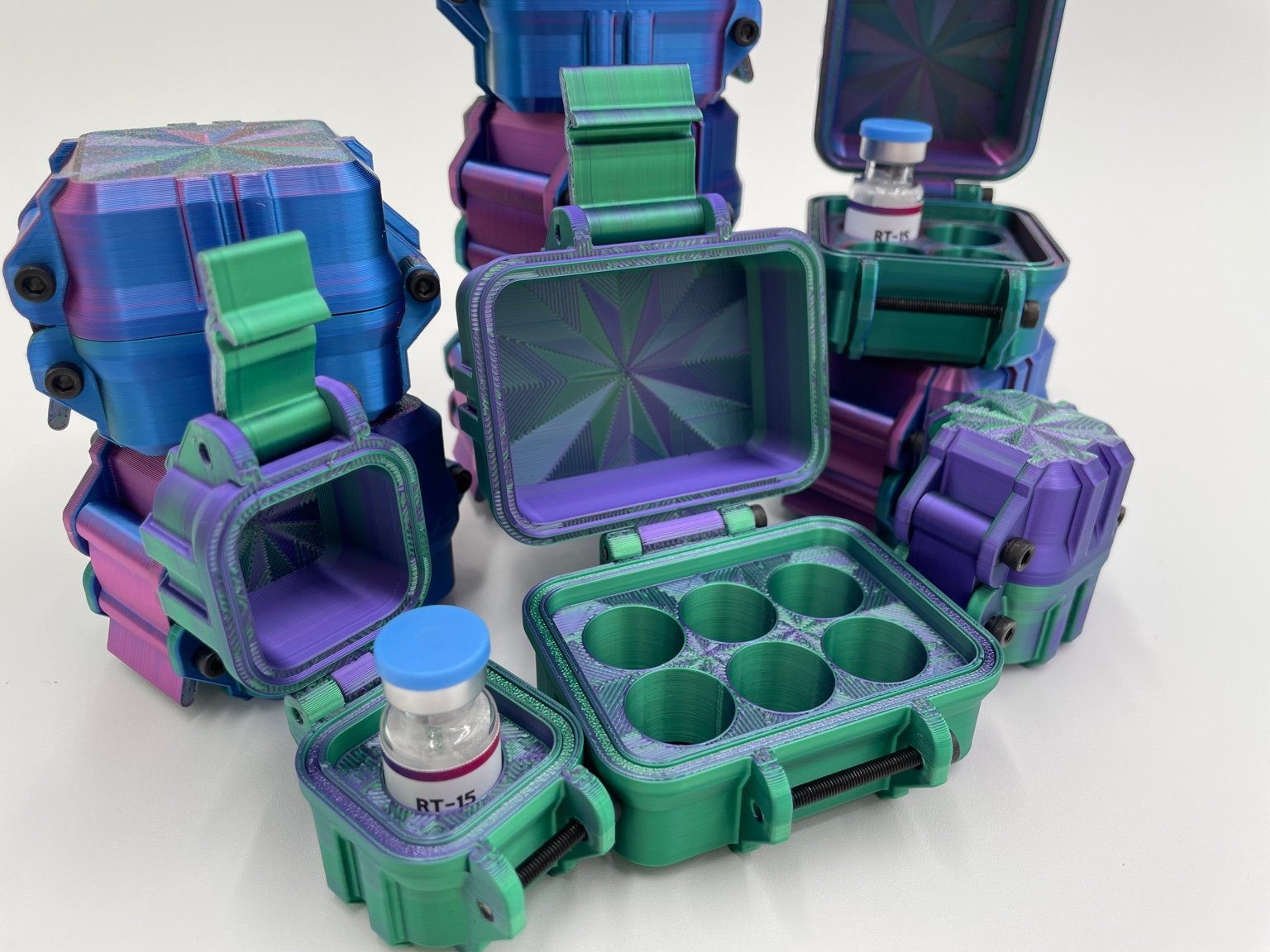 A collection of colorful, multi-colored cases in shades of green, purple, and blue. Some cases are open and show a vial labeled 'Peptide Basix RT-15' inside, while others are empty or have a compartment with spaces for vials.